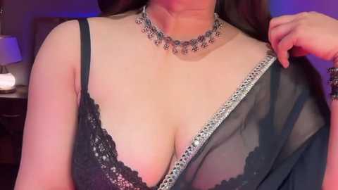 Video of a woman in a black lace bra and sheer black robe, adorned with a silver choker necklace. She has a fair complexion, and the background is dimly lit with purple and white lighting.