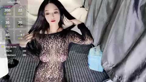 Video of an East Asian woman with long black hair, wearing a sheer, black leopard print bodysuit, sitting on a striped bed in a dimly lit room, with a virtual reality headset displaying a temperature gauge.