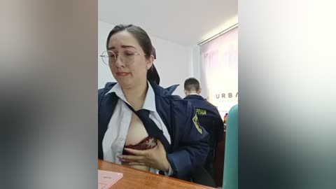 A video shows a Caucasian woman with fair skin and glasses, wearing a partially unbuttoned white shirt exposing her breasts, seated at a desk in an office setting.