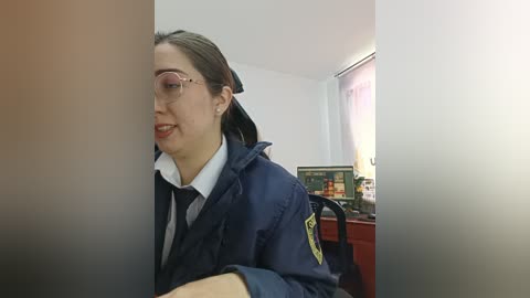 Video of a young Asian woman in a navy blue police uniform, seated indoors, with a white shirt and glasses, in a dimly lit room with a computer and red furniture.