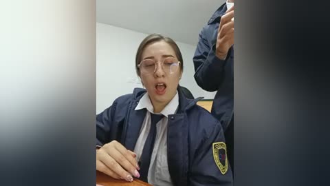 Video of a female police officer with light skin, glasses, and short brown hair, wearing a navy uniform and tie, appearing distressed, holding a phone in a dimly lit room.
