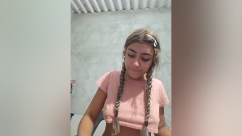 Video of a young woman with light skin and braided hair, wearing a pink crop top, standing in a concrete-walled room.