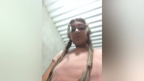 A blurry video of a topless woman with long, blonde braided hair, standing in a narrow hallway with white blinds covering a window. Her skin tone is light, and she appears to be of Caucasian ethnicity.
