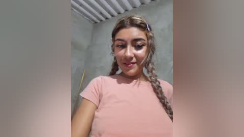 Video of a young woman with light brown hair in braids, wearing a pink t-shirt, standing in a dimly lit room with grey walls and white ceiling tiles.