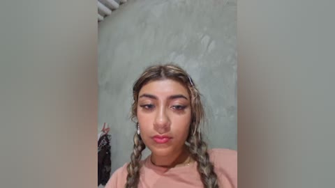 Video of a young woman with light brown skin, styled with braided hair, wearing a pink top. Background shows a textured, muted green wall and a glimpse of a black dress.