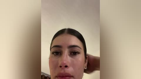 A video of a close-up of a young woman with fair skin and dark hair, slightly parted. She has a neutral expression, wearing small earrings, with a man's hand gently touching her neck.