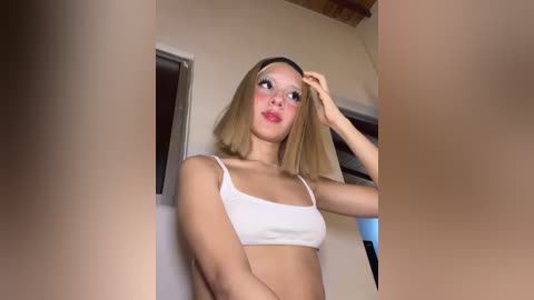 Video of a young, light-skinned woman with straight, shoulder-length blonde hair, wearing a white crop top and standing in a dimly lit room.