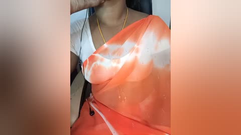 A video of a woman in a white and orange saree, with a wet, see-through effect, wearing a gold necklace, her face partially visible. The background is blurred.
