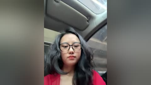 Video of an Asian woman with long, wavy black hair and glasses, wearing a red top, seated in a car with a grey interior.