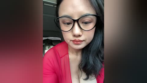 Video of an Asian woman with medium skin tone, wearing black-rimmed glasses, a red cardigan, and light makeup. She has long black hair, and is in a car, with blurry background.