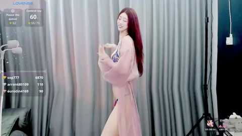 Video of a smiling, slender, red-haired woman with large breasts, wearing a sheer pink robe, standing confidently in a dimly lit room with grey curtains and a camera setup.