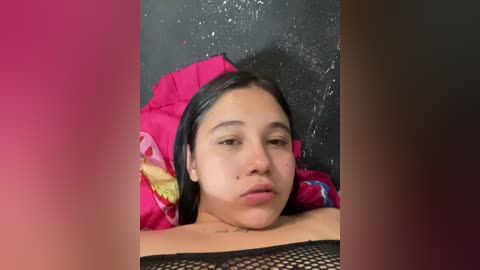 Video of a young Latina woman with medium skin tone, lying on a colorful pillow against a dark wall, wearing a black mesh top. Her expression is neutral, and her straight black hair is slightly wet.