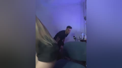 A blurred video shows a young man in a black T-shirt and shorts, with long blonde hair, bending over a blue couch. The room is dimly lit with blue lighting, and a white mug and bottle are visible on a nearby table.