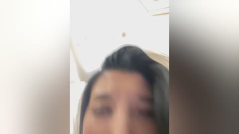 Blurry video of a person with black hair, focusing on the top of their head and part of their face, in a brightly lit room with a white ceiling and a white wall.