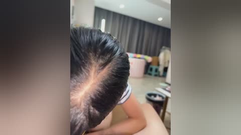 A close-up video of a person with dark hair, partially hidden by a large, curly wig. The background features a modern, minimalist room with beige walls, dark curtains, and a cluttered desk.