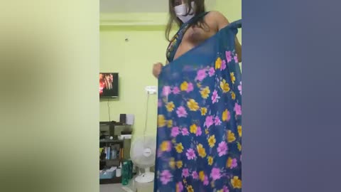 A video of a woman with medium skin tone, wearing a floral-patterned blue blanket, standing in a dimly lit room with a TV and shelves visible in the background.