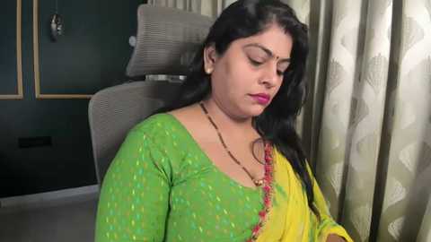 Video of a South Asian woman in a green and yellow sari, seated on a grey chair, with a contemplative expression.