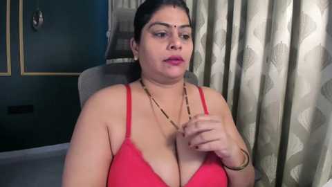 Video of a South Asian woman with medium skin tone, wearing a red bra, adjusting her cleavage, in a dimly-lit room with patterned curtains.