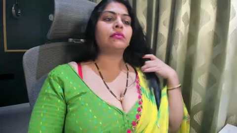 Video of a South Asian woman with long black hair, wearing a green blouse with red accents, sitting in a modern chair.