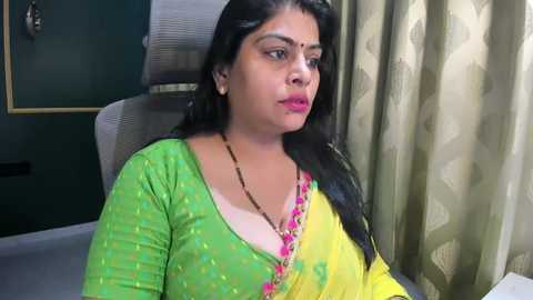 Video of a South Asian woman with long black hair, wearing a green and yellow sari, sitting on a gray chair, in a room with dark green walls and beige curtains.