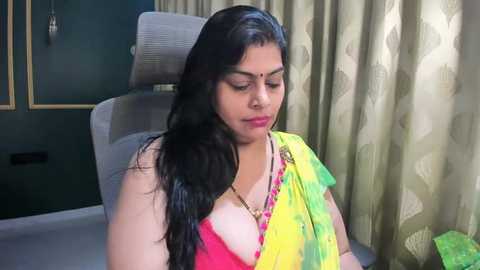Media: Video of a South Asian woman with long black hair, wearing a pink bra and yellow-green sari, seated on a grey chair in a dimly lit room with green curtains.