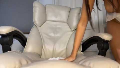 A video of a nude woman with long brown hair, light skin, and medium breasts, sitting on a white leather recliner, adjusting her position.