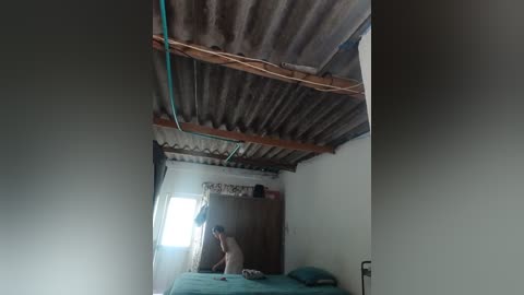 Video of a small, dimly lit room with corrugated metal roof and walls, featuring a person wearing a white shirt and blue pants, standing near a bed with a green sheet.