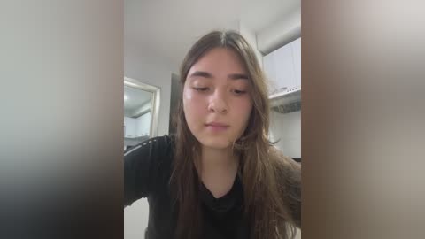 A video of a young woman with long, straight brown hair, wearing a black top, standing in a modern, white kitchen with visible counters and cabinets.
