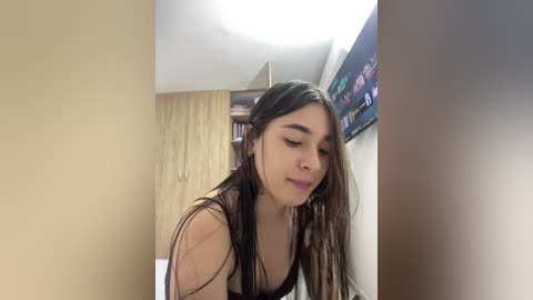 Video of a young woman with long dark hair, wearing a black bra, in a dimly lit bedroom with beige walls, a wooden wardrobe, and a TV displaying colorful images on the right.