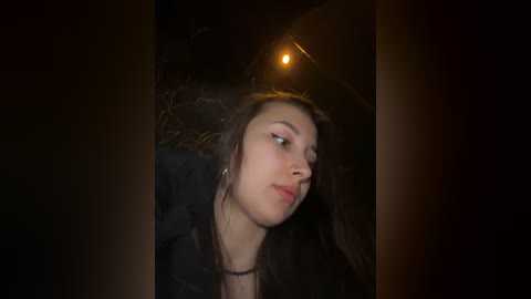 Video of a young woman with long brown hair, wearing a black jacket, looking contemplative, taken at night with dim streetlight in the background.