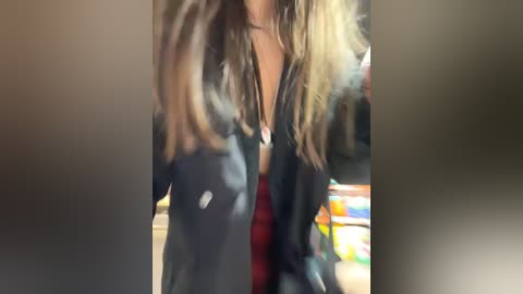 Video of a woman with long, wavy, light brown hair, wearing a black leather jacket, red dress, and a necklace, standing indoors. Background shows blurred store shelves with colorful products.