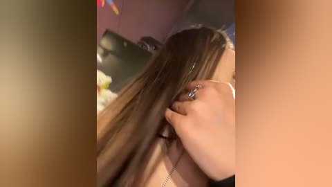 Media: Video of a woman with long, straight brown hair, being styled by another person, wearing a beige top, in a dimly lit indoor setting with blurred background.