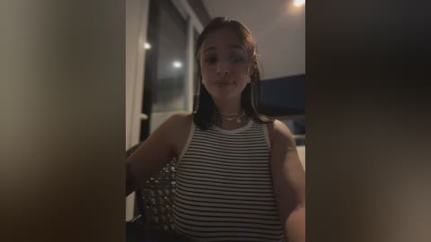 Video of a young woman with medium skin tone, straight dark hair, and a sleeveless striped top, standing indoors. She wears a gold necklace and is in a dimly lit room with blurred background elements.