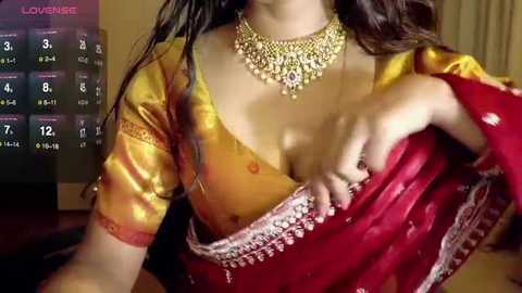 A video of a woman in a red and gold saree, with a gold necklace, partially revealing her breasts, in a dimly lit room with a calendar on the left.
