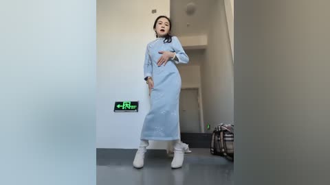 A video of an Asian woman in a light blue medical gown and white shoes, standing in a modern, minimalist hallway with a green exit sign and a brown bag on the floor.