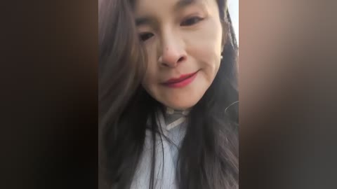 Video of a young woman with long, dark hair, fair skin, and a closed-eye smile. She wears a light gray shirt, standing in front of a blurred, dark background.