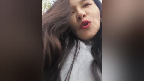 A video of a young Asian woman with long black hair, fair skin, and a white turtleneck sweater, puckering her lips in a playful manner. The background is blurred, with soft, natural lighting.
