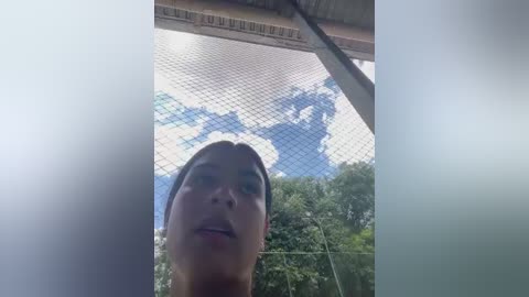 Video of a young woman with light skin, brown hair, and a neutral expression, standing under a chain-link fence in a green, leafy outdoor setting with a partly cloudy sky.