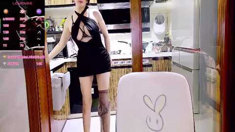 Video of a woman in a revealing black dress with crisscross straps, standing in a modern kitchen with a Playboy chair.