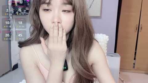 Video of an Asian woman with long, wavy brown hair, wearing a pink spaghetti strap top, covering her mouth in a shy or embarrassed gesture. Background shows a purple wall, white furniture, and a wooden door.