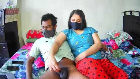 Video of a Black man and woman lying on a colorful bed, wearing masks, in a messy bedroom.