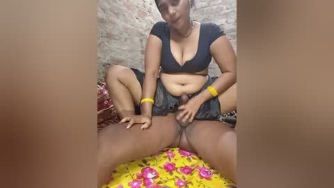 A video showing a South Asian woman with medium skin tone, wearing a black crop top, sitting on a yellow bedspread with pink flowers. She is spreading her legs, revealing a partially erect penis. The background features a brick wall.
