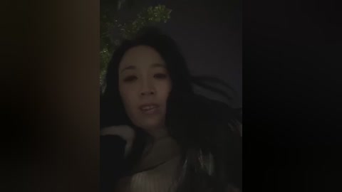 A dimly lit video of an Asian woman with long black hair adorned with a floral crown, wearing a dark, shiny jacket. Her expression is serene, eyes closed, set against a dark, blurry background.