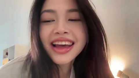 A close-up video of a smiling young woman with long, straight dark hair, light skin, and closed eyes. She wears a white top. The background features a beige wall with a warm light.