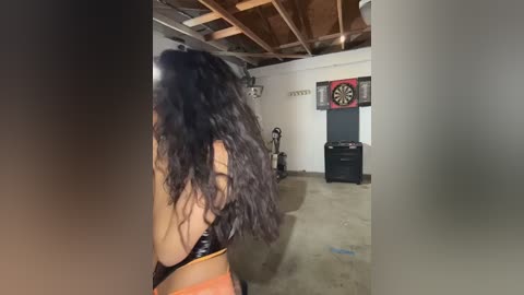Video of a woman with long, curly black hair, wearing a black and orange sports bra, standing in a dimly lit, unfinished basement with exposed wooden beams, dartboard, and a dartboard cabinet.
