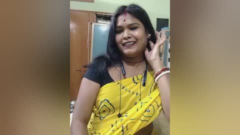 Video of an Indian woman with long black hair, wearing a yellow sari with black patterns and a black top, smiling in a kitchen with wooden cabinets and a stainless steel fridge in the background.