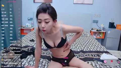 A video of an Asian woman in black lingerie, adjusting her bra on a bed with patterned sheets in a messy bedroom.