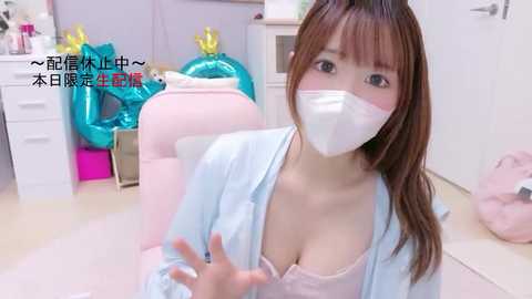 Video of a young Asian woman in a white surgical mask, light blue robe, and a revealing pink bra, sitting on a pink chair in a cluttered room with blue balloons and a white dresser.