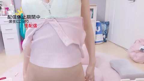 Video of a woman in a pastel pink wrap top and beige skirt, standing in a tidy, light-colored room with white furniture and a pink cushion on the floor.