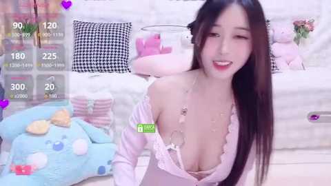Media: Video of an Asian woman with long black hair, wearing a low-cut, pastel pink off-shoulder dress, smiling, surrounded by colorful pillows, plush toys, and a video recording overlay.